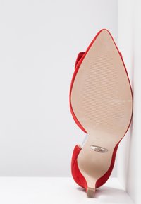 Chaussure à talon pointu en daim rouge, positionnée semelle vers le haut contre un coin blanc, montrant la semelle texturée beige et un petit talon kitten.