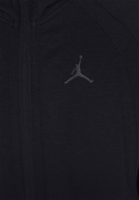 Černá sportovní bunda z hladkého materiálu, s zipem, vysokým límcem a šedým logem Jumpman na levé části hrudi.