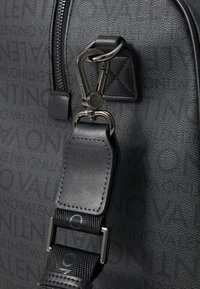 Saco de tecido preto com uma superfície texturizada, apresentando um fecho metálico e uma alça de couro. O design notável inclui detalhes em relevo do logótipo.