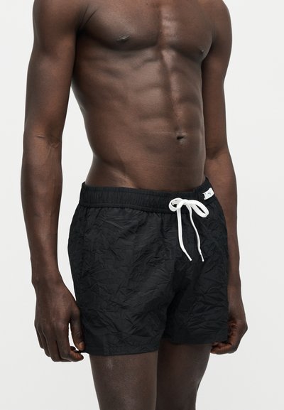 Αθλητικός άνδρας φοράει μαύρο ζαρωμένο μαγιό με λευκό κορδόνι, εμφανιζόμενος από το μέσο του στήθους μέχρι το μέσο του μηρού σε απλό φόντο.