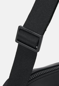 Czarny pasek z tkaniny z prostokątną czarną klamrą, z widoczną teksturą i trwałym szyciem, przymocowany do torby.