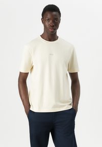 Cream short-sleeve T-shirt with a round neckline, soft fabric, and a subtle "BOSS" logo at the center. Paired with navy trousers.