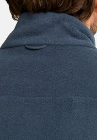Marineblaue Fleecejacke, weiche Textur, mit hohem Kragen und einem kleinen Schlaufen-Detail auf der Rückseite. Nahtnähte sind über das Material sichtbar.