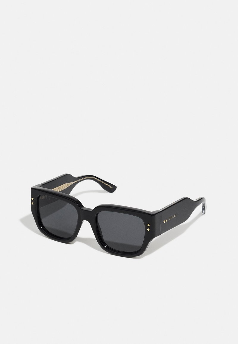 Black oversized sunglasses with square lenses, featuring gold accents and a smooth texture. Designed with thick frames and subtle brand detailing.