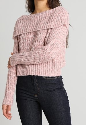Femme portant un pull épaissi rose tricoté épaules dénudées à manches longues, associé à un jean bleu foncé.
