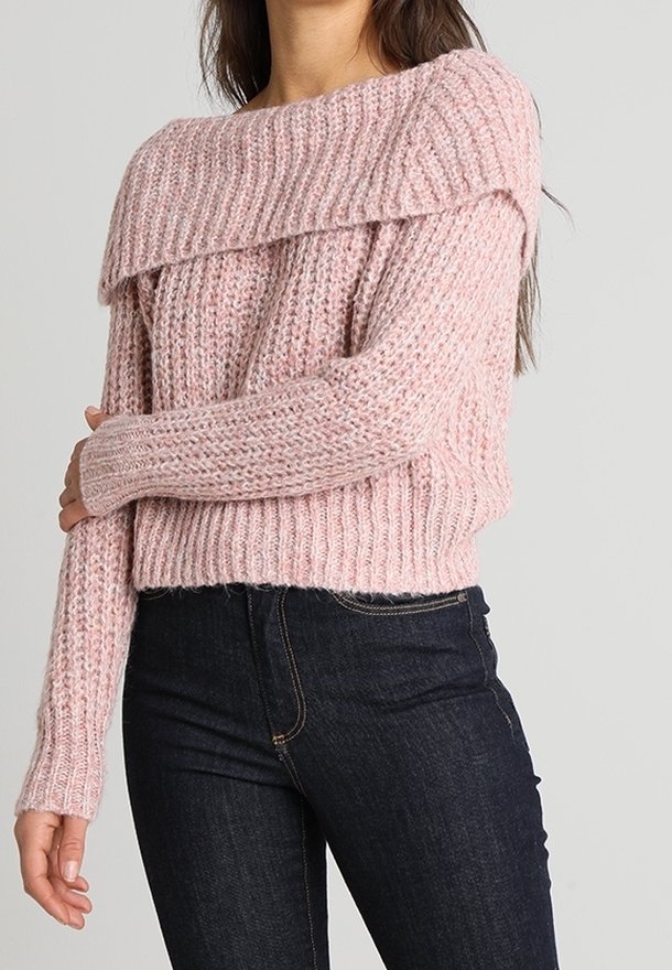 Femme portant un pull épaissi rose tricoté épaules dénudées à manches longues, associé à un jean bleu foncé.