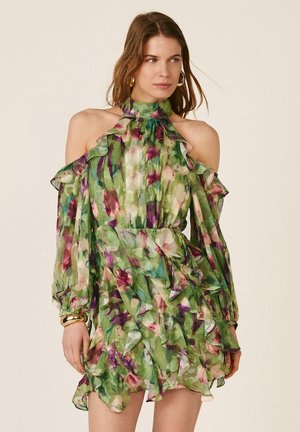 Mujer con un vestido verde con flores, estilo cold-shoulder con volantes y mangas largas transparentes, complementado con pendientes de aro dorados y pulseras.