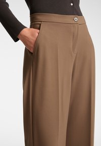 Person som bär bruna skräddarsydda byxor med knappstängning och handen i höger ficka, i kombination med en svart långärmad topp.