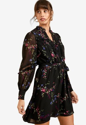 Schwarzes Blumenkleid mit langen durchsichtigen Ärmeln, das eine geraffte Taille, Spitzenakzente und farbenfrohe Blumenmuster in Pink, Blau und Grün aufweist.