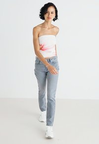 Hvit stroppeløs topp med rosa blomsterprint, kombinert med lyseblå denimjeans og hvite joggesko, båret av en person som går mot en ensfarget bakgrunn.