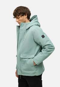 Chaqueta verde menta con capucha, cierre frontal de cremallera, dos grandes bolsillos frontales y un parche en la manga. Textura suave con un corte moderno.