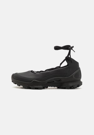 Chaussure de sport noire avec semelle robuste, détails découpés et lien à la cheville, vue de profil sur fond clair.