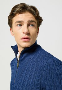Granatowy sweter z zamkiem błyskawicznym, wykonany z fakturowanego splotu w kablowym wzorze, z złotym zamkiem i ściągaczem, podkreślający przyjemny materiał.