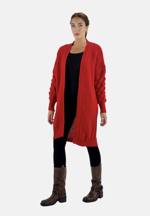 Cárdigan de punto rojo con un frente abierto, mangas abullonadas texturizadas y un corte relajado, combinado con leggings negros y botas de tobillo marrones.