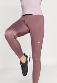 Leggings de Nike en un tono malva, hechos de un material suave, con un diseño ajustado y un pequeño logo blanco. Combinados con zapatillas deportivas negras.
