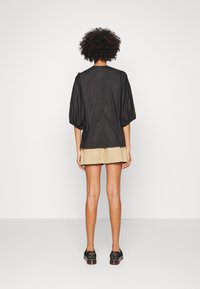 Persona con capelli ricci che indossa una maglietta nera a maniche a sbuffo, pantaloni beige e scarpe nere, in piedi di fronte a un muro bianco.