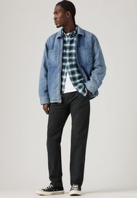 Lysblå denimjakke, parret med en grønternet skjorte, hvid t-shirt og mørke bukser. Sorte sneakers fuldender outfittet.