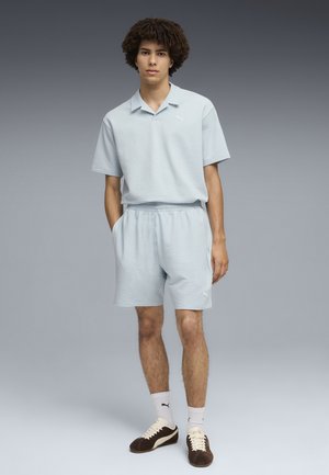 Joven de pie con camisa de manga corta texturizada azul claro y pantalones cortos a juego, calcetines blancos Puma y zapatillas marrones Puma.