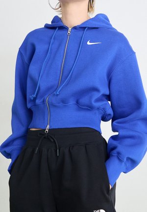Persoon die een blauwe cropped Nike hoodie met rits en trekkoorden draagt en zwarte The North Face joggingbroek met handen in de zakken.