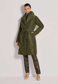 Olijfgroene gewatteerde jas met een brede kraag, ceintuur in de taille en lange mouwen. Gepaard met gestructureerde slangenprint broek en beige hakken.