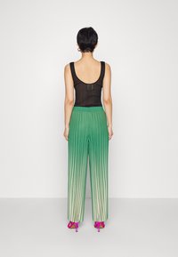 Eine Person steht mit dem Rücken zur Kamera und trägt ein schwarzes ärmelloses Oberteil, grüne plissierte Ombre-Hosen und rosa High-Heels.