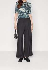 Modell som bär en åtsittande topp i teal och svart med abstrakt mönster och korta ärmar, parat med hög midja och vida svarta byxor samt svarta loafers.