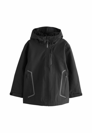 Person mit lockigem Haar, die eine schwarze Kapuzenjacke trägt, die bis zum Kinn hochgezogen ist, blickt links vor schlichtem Hintergrund.