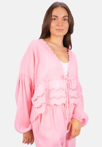 Helles rosa, kurz geschnittenes Blusenshirt aus weicher Baumwolle, mit gewelltem Spitzenbesatz, lockeren Ärmeln und einem Krawattenverschluss vorne, über einem weißen Oberteil getragen.