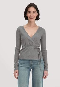 Lächelnde Frau mit kurzem dunklem Haar, die einen grauen Wickel-Pullover mit bindegürtel und hellblauen Jeans vor einem schlichten Hintergrund trägt.