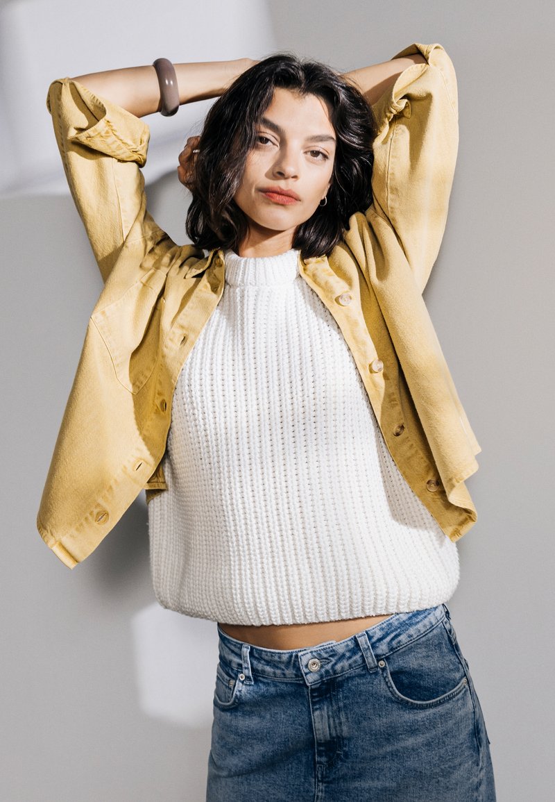 Camicia gialla con bottoni sopra un maglione bianco lavorato a maglia, abbinata a jeans in denim blu a vita alta. Tessuti morbidi e vestibilità casual.