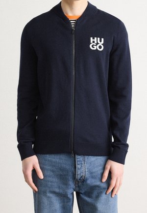 Man draagt een marineblauwe trui met rits en een wit "HUGO"-logo op de linkerborst, een gestreept overhemd eronder en een blauwe spijkerbroek.