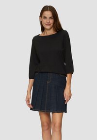 Zwarte blouse met lange mouwen, gecombineerd met een donkere spijkerrok in A-lijn met plooien en contrasterende stiksels, getoond op een neutrale achtergrond.