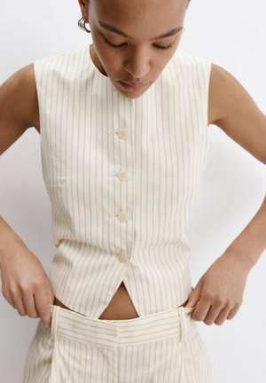 Femme portant un haut beige rayé sans manches à boutons, ajustant un pantalon rayé taille haute assorti sur fond blanc.