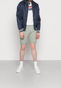 Granatowa satynowa kurtka bomberka z białymi paskami przy mankietach, biała koszulka z logo, jasnooliwkowe szorty i białe sneakersy. Gładkie faktury, dopasowany krój.