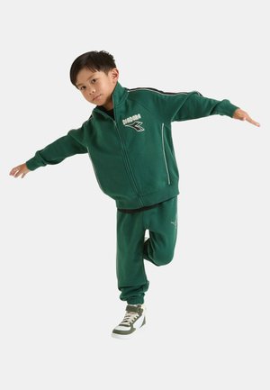 Tuta verde con giacca con zip e pantaloni. Dettagli in bianco e logo. Modello posa in modo dinamico con scarpe da ginnastica alte.