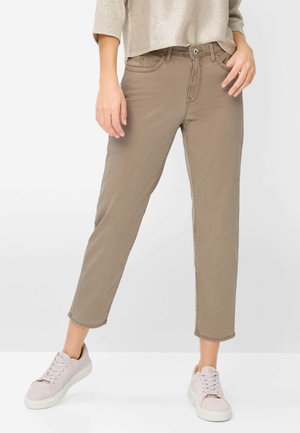 Femme portant un pantalon beige cropé à jambes droites, un pull léger en tricot, et des baskets rose pâle, debout devant un fond neutre.
