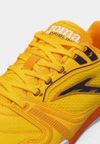 Φωτεινό κίτρινο αθλητικό παπούτσι με πορτοκαλί και μαύρες λεπτομέρειες, κίτρινες κορδόνια, επάνω μέρος από διαπνέον δίχτυ και λογότυπο "Joma Dribling" στη γλώσσα.