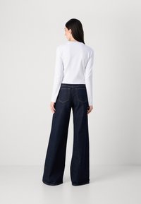 Modella di spalle, che indossa una camicia bianca a maniche lunghe e jeans a gamba larga in denim scuro, su uno sfondo chiaro e semplice.