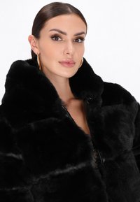 Cappotto in pelliccia sintetica nera con colletto alto, caratterizzato da una superficie texture e grandi bottoni rotondi. Accentuato da una fodera liscia.