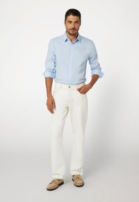 Šviesiai mėlynas ilgos rankovės marškinėliai su mygtukų apykakle, derinami su baltais kelnais. Modelis dėvi smėlio spalvos slip-on batus. Švarus, modernus siluetas.