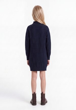 Personne aux longs cheveux blonds portant une robe en maille bleu marine à manches longues et des bottines marron, debout de dos sur fond blanc.