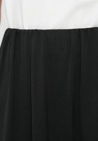 Jupe noire avec une taille froncée et une texture lisse. Le haut est en tissu blanc uni, créant un look bicolore.