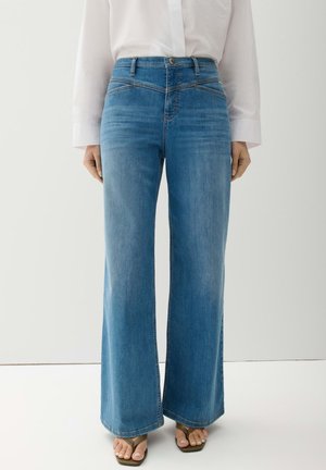 Persoon draagt hooggesneden blauw wijd uitlopende spijkerbroek met een wit shirt met lange mouwen en bruine sandalen met open tenen.