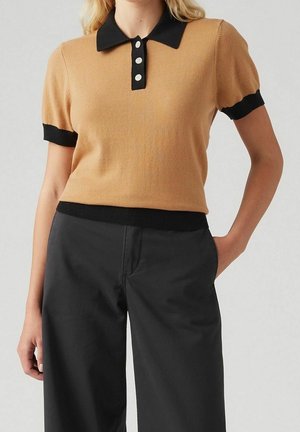 Femme portant un polo à manches courtes couleur camel avec col et poignets noirs, associé à un pantalon noir taille haute, debout avec une main dans la poche.