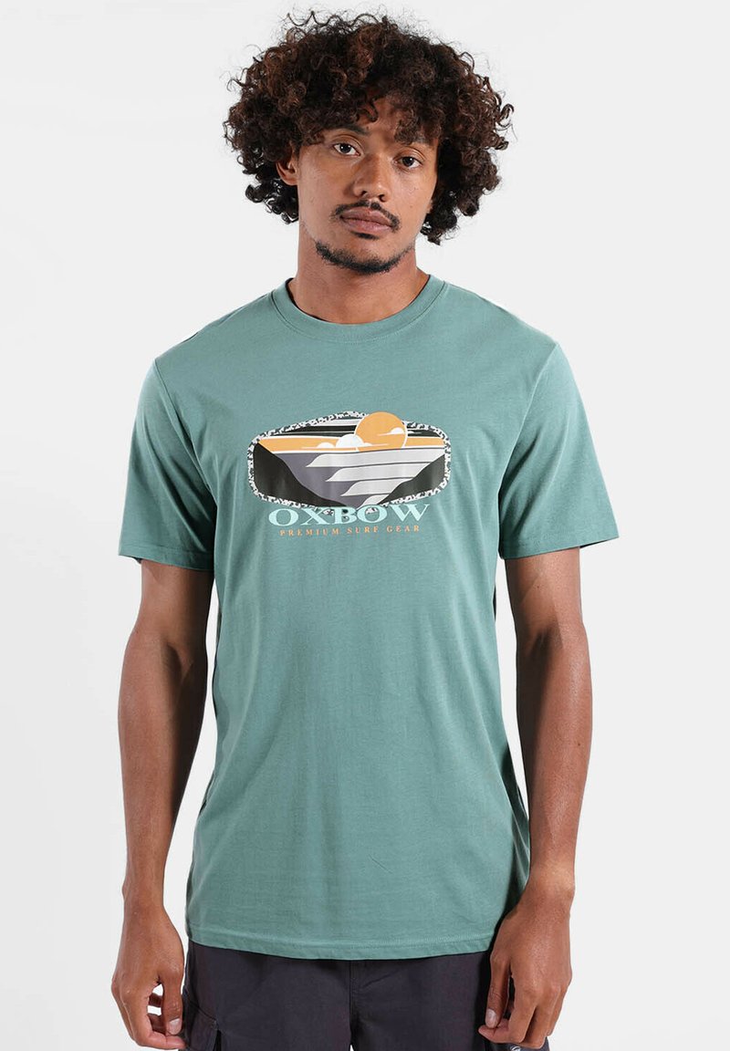 T-shirt à manches courtes couleur sarcelle avec un motif graphique représentant un coucher de soleil et des vagues. Confectionné en tissu doux, avec un col rond et une coupe décontractée.
