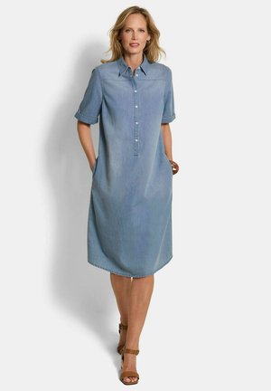 Frau läuft und trägt ein knieumspieltes hellblaues Jeanskleid mit kurzen Ärmeln und braunen Sandalen mit Knöchelriemen, die Hände in den Taschen des Kleides.