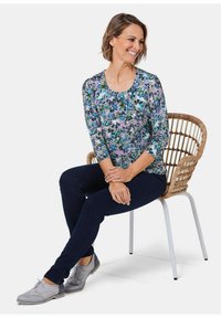 Bluse mit Blumenmuster und Dreiviertelärmeln, in verschiedenen Blau- und Grüntönen auf einem hellen Hintergrund. Kombiniert mit dunklen Jeans und grauen Schuhen.