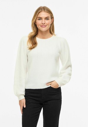 Fiatal nő szőke hajjal, fehér bordázott pulóvert és fekete nadrágot viselve, egy kézzel a zsebében áll fehér háttér előtt.