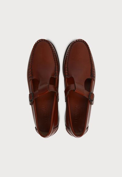 Paire de mocassins marron en cuir poli à enfiler avec des sangles à boucle et des coutures visibles sur fond blanc uni.
