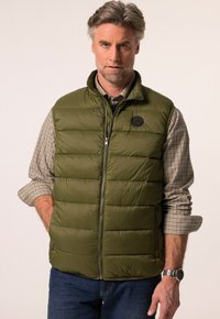 Zelená vyp padded vesta se zipem vpředu a kulatým logem na hrudi, nošená přes kostkovanou košili. Má boční kapsy a vysoký límec.